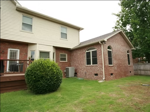 Backyard of a two-story house. Brick extension with arched windows and a deck. Green grass and shrubbery present.