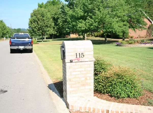 Brick mailbox with the number 115, on a green lawn, with a truck parked on the road, and trees in the background.