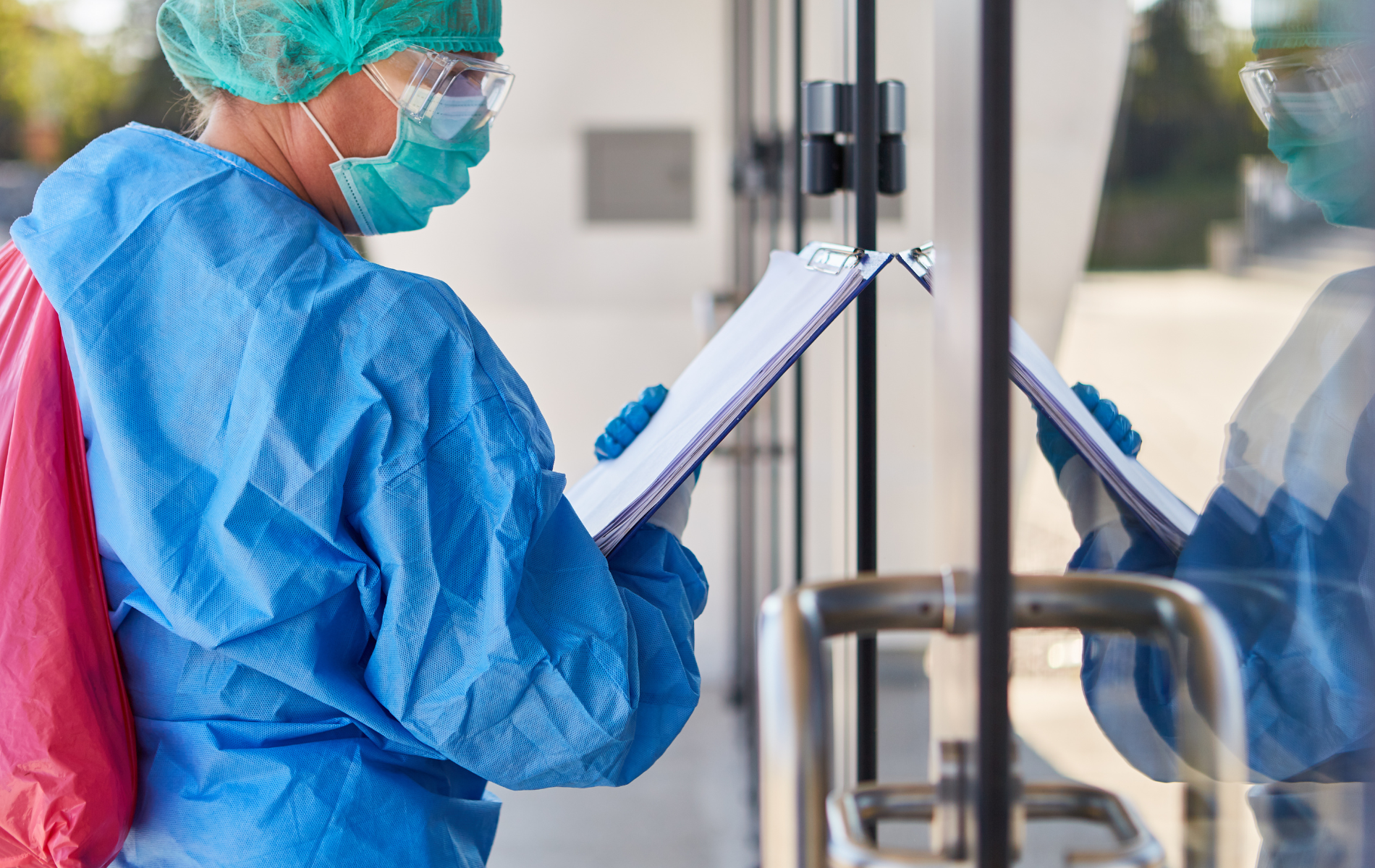 A nurse in a protective suit is standing in front of a door holding a clipboard.