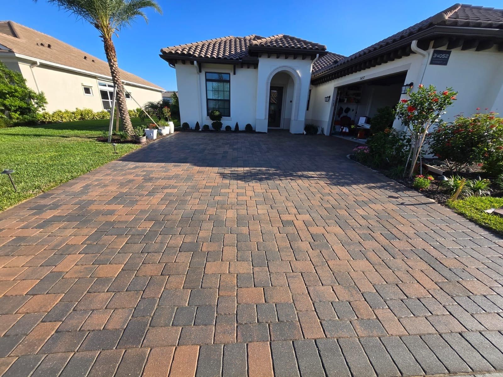 Brick paved driveway leading to a house with a white exterior, garage, and brown tiled roof on a sunny day.