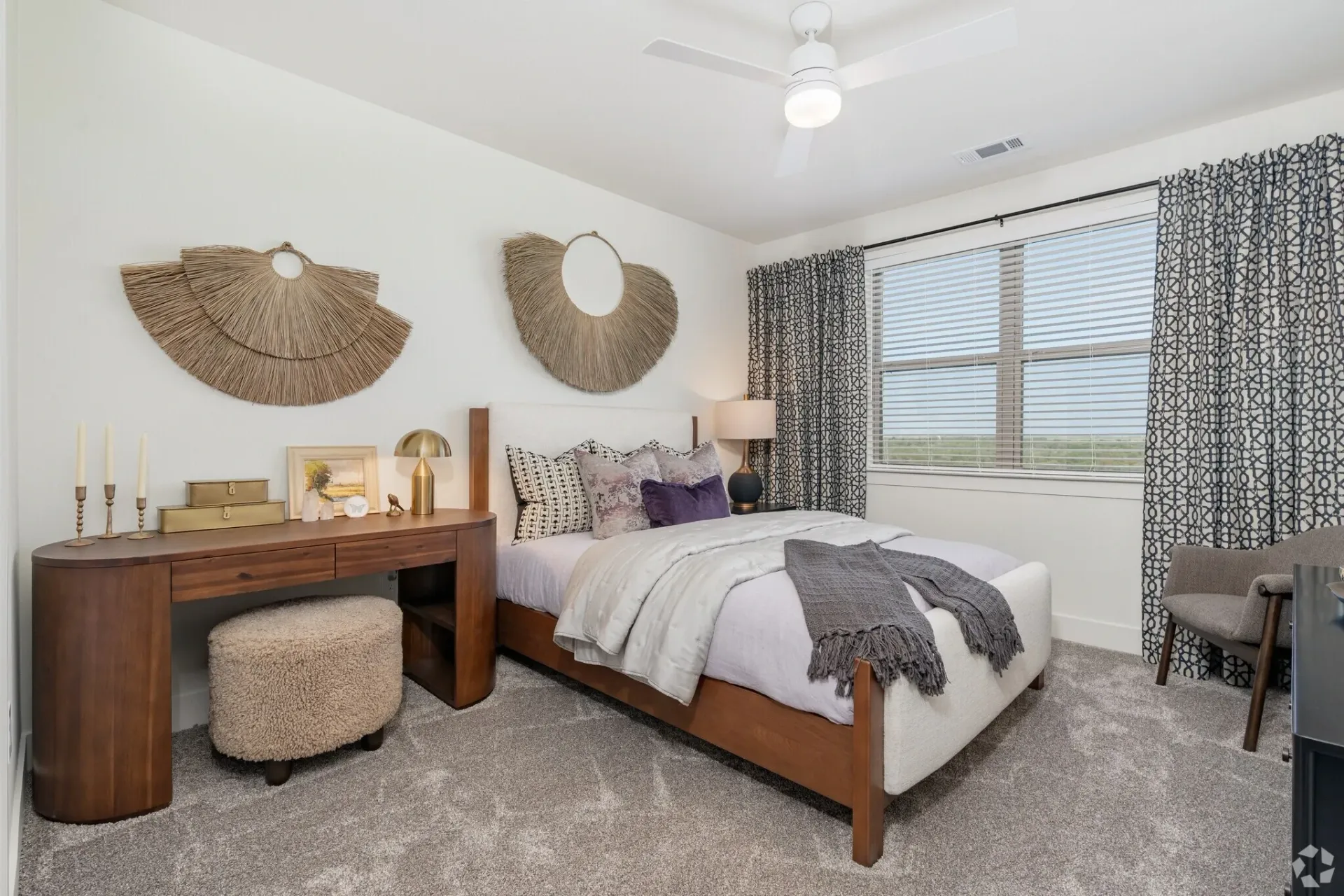 A well-lit bedroom with a wooden bed, a vanity with a textured stool, two wall decorations, and patterned curtains.
