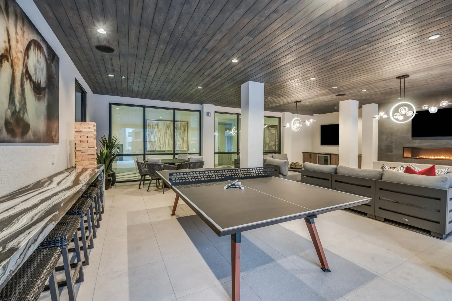 A modern recreation room with a ping pong table, lounge seating, a bar counter, and a dark wooden paneled ceiling.
