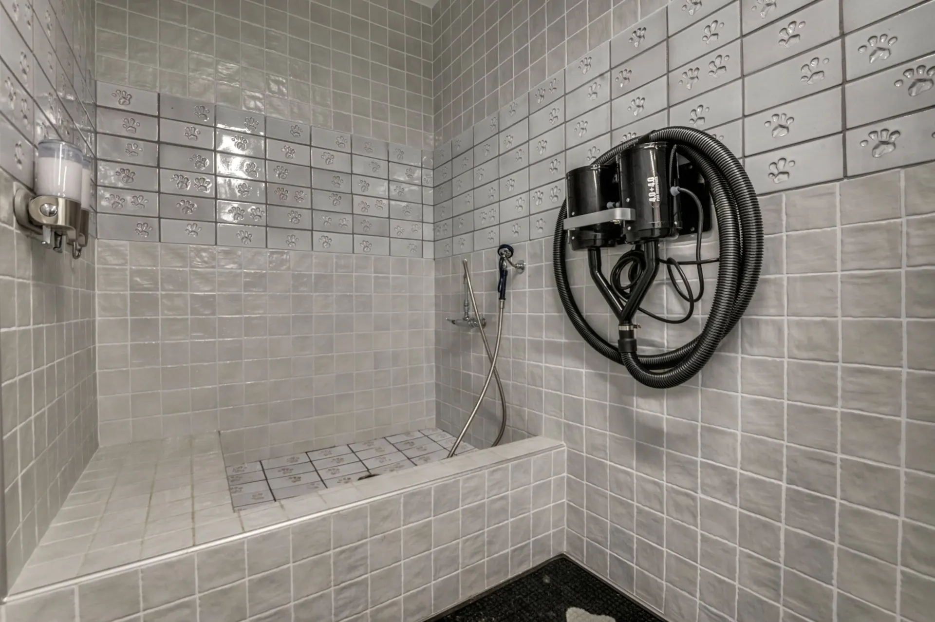 Pet grooming station with raised tub, shower head, and powerful dryers. Tiled walls feature paw print accents.
