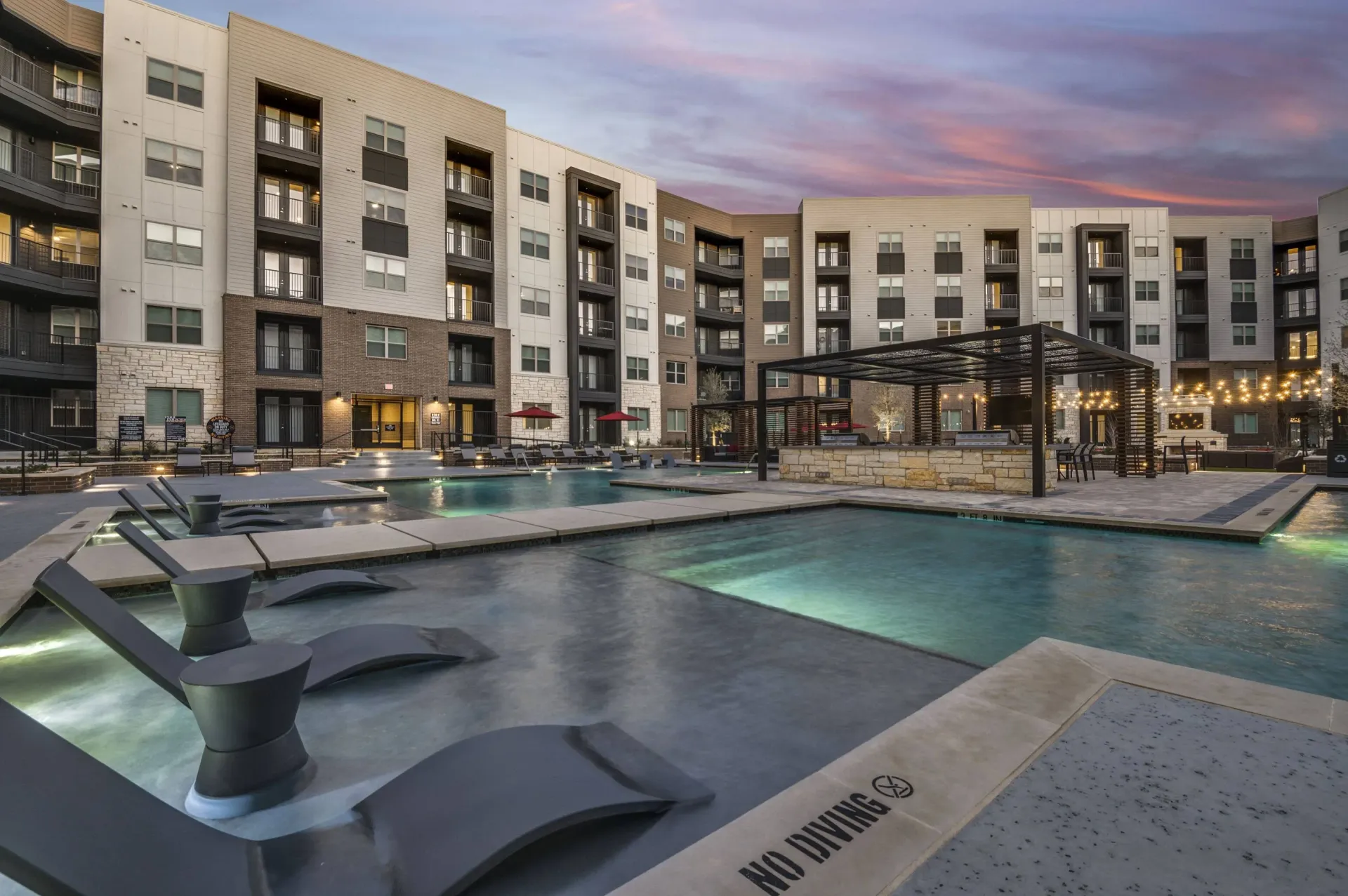 Modern apartment complex pool area with lounge chairs and outdoor seating at dusk.