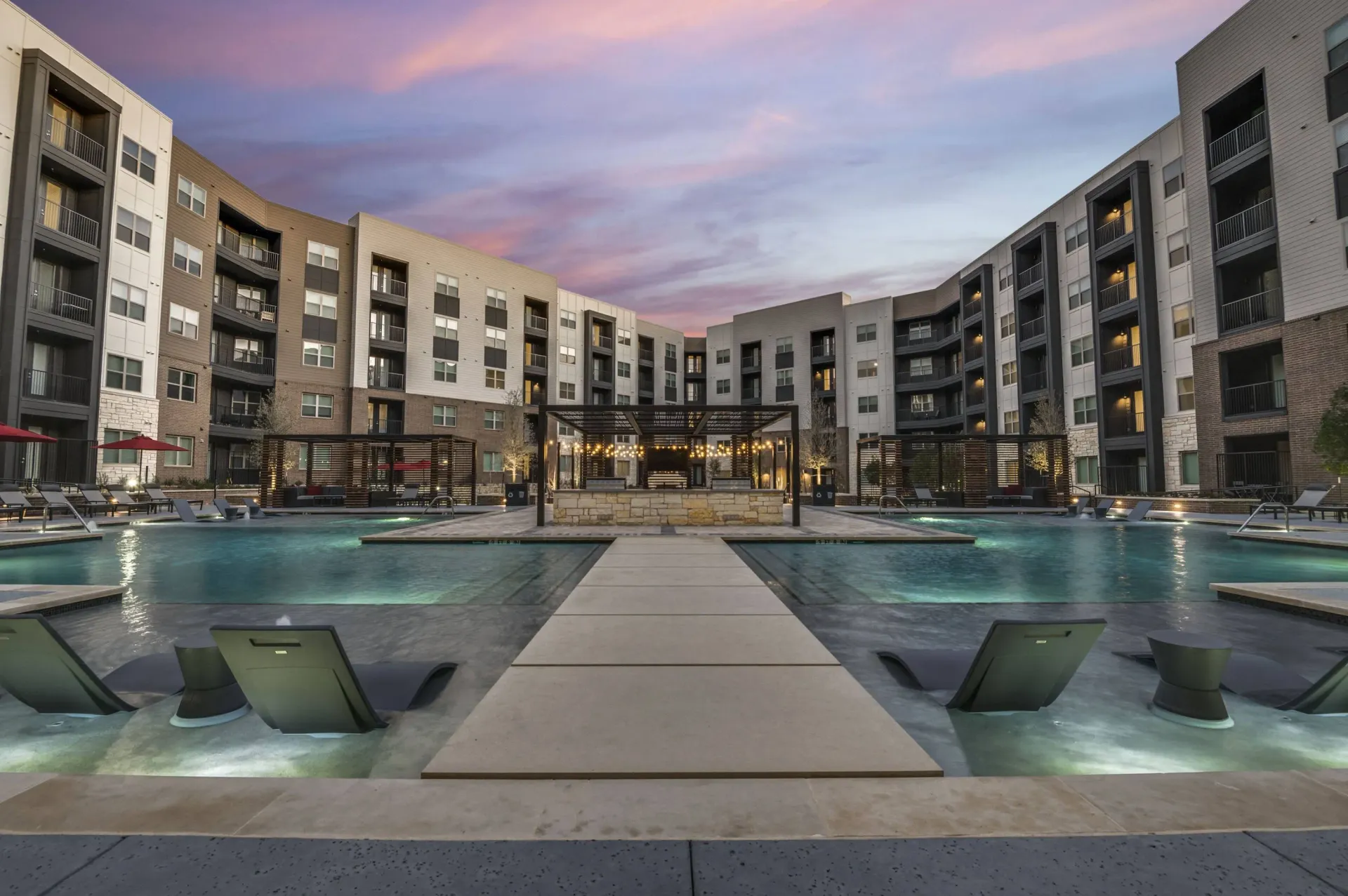 Luxurious resort-style pool area with lounge chairs and modern apartment buildings at dusk.