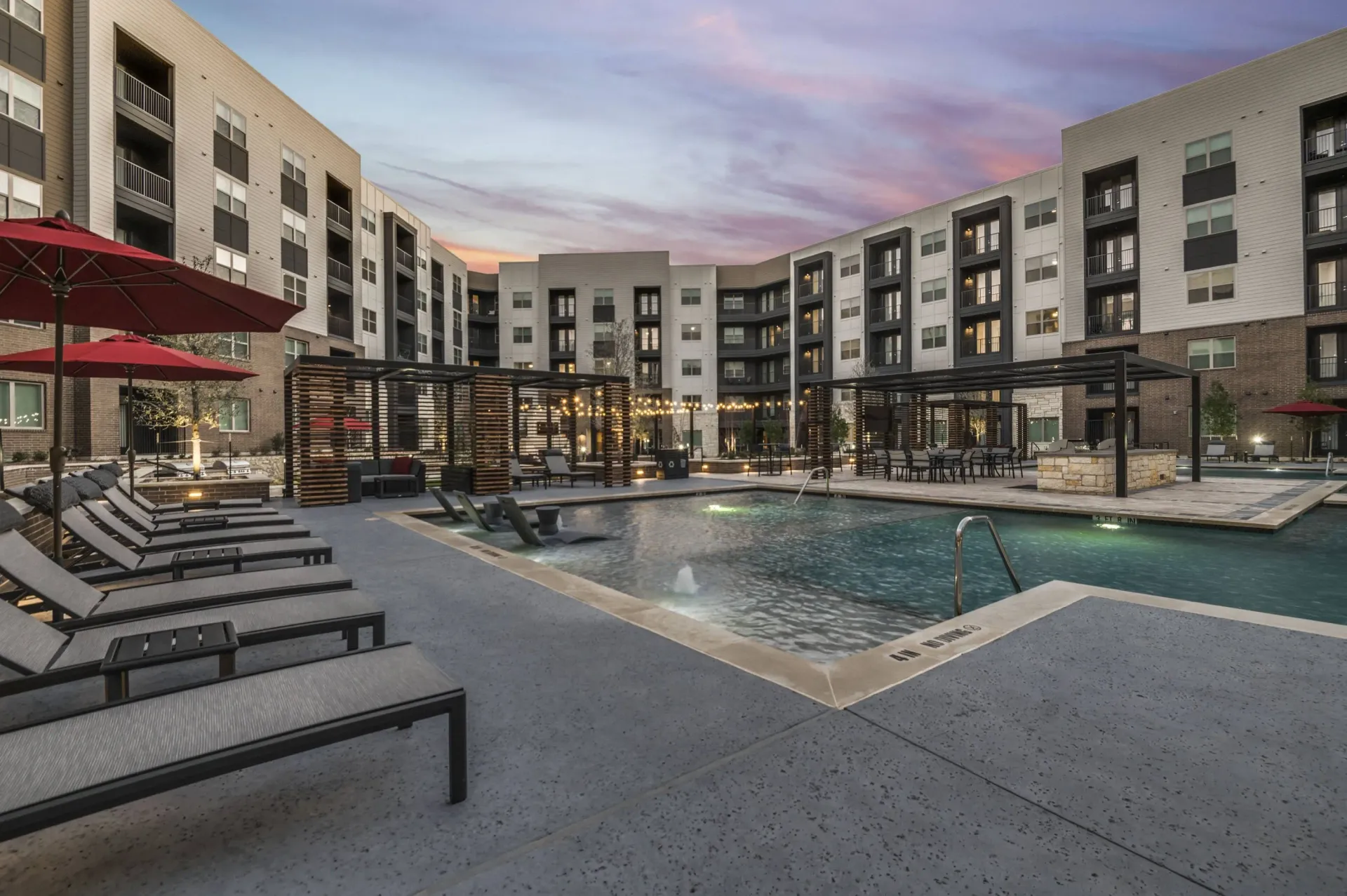 Luxury apartment complex courtyard with a swimming pool, lounge chairs, and covered seating areas at dusk.