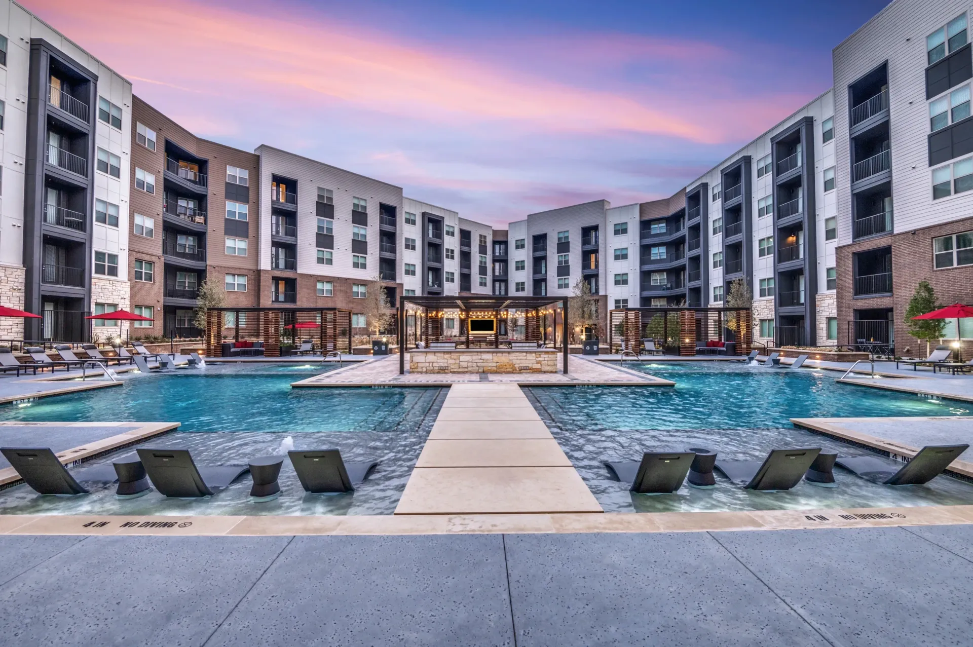Resort-style pool with lounge chairs and cabanas at an apartment complex during sunset.