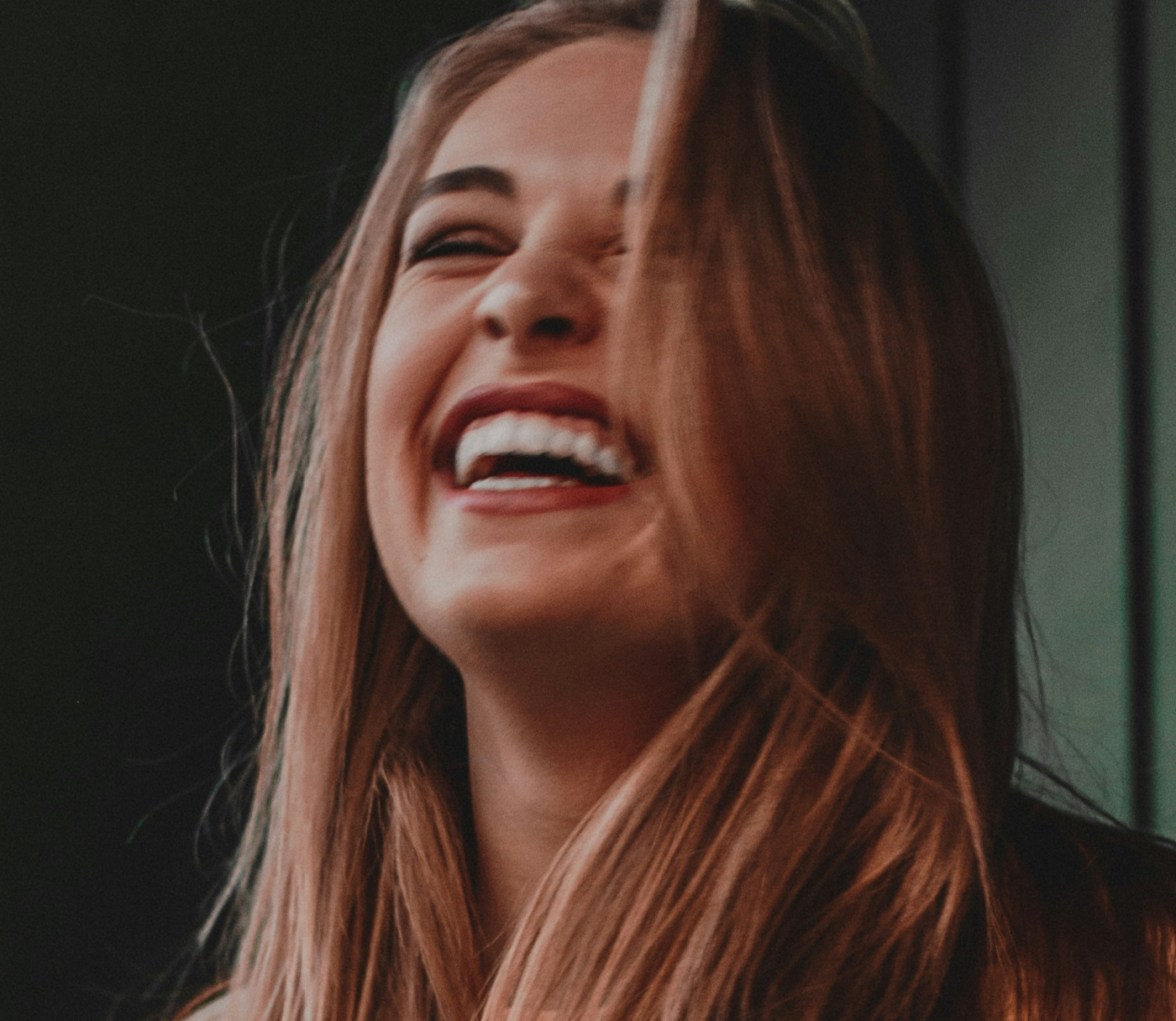 Smiling woman laughing, with long brown hair, in soft indoor light