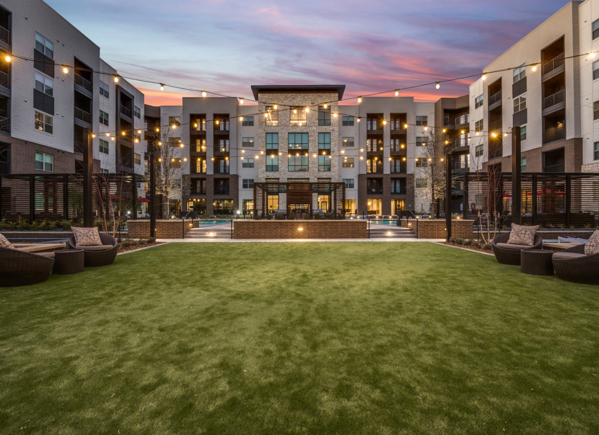 Modern apartment courtyard with green lawn, lounge seating, and lit buildings at dusk