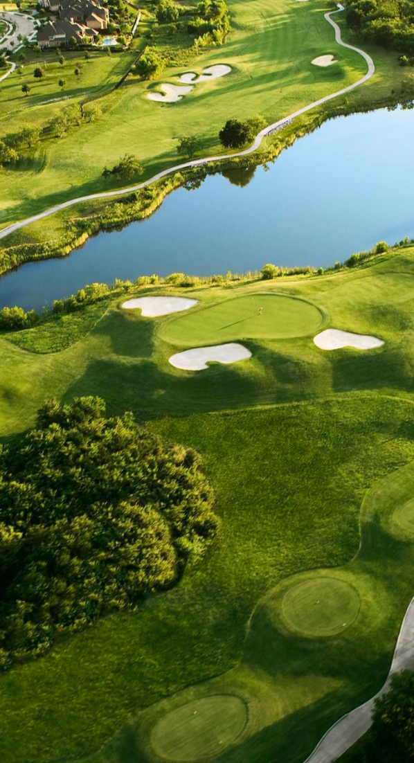 An aerial view of a green golf course with sand bunkers and a winding water hazard under bright sunlight.