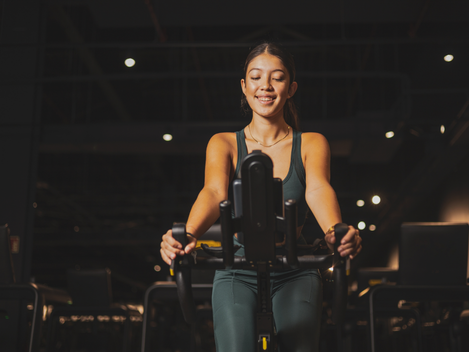 A person in dark green athletic wear smiles while riding a stationary exercise bike in a dim, modern gym.
