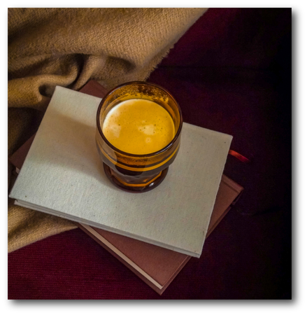 A glass of golden coffee sits atop a pale book, resting on a darker book and a tan blanket against a deep purple backdrop.