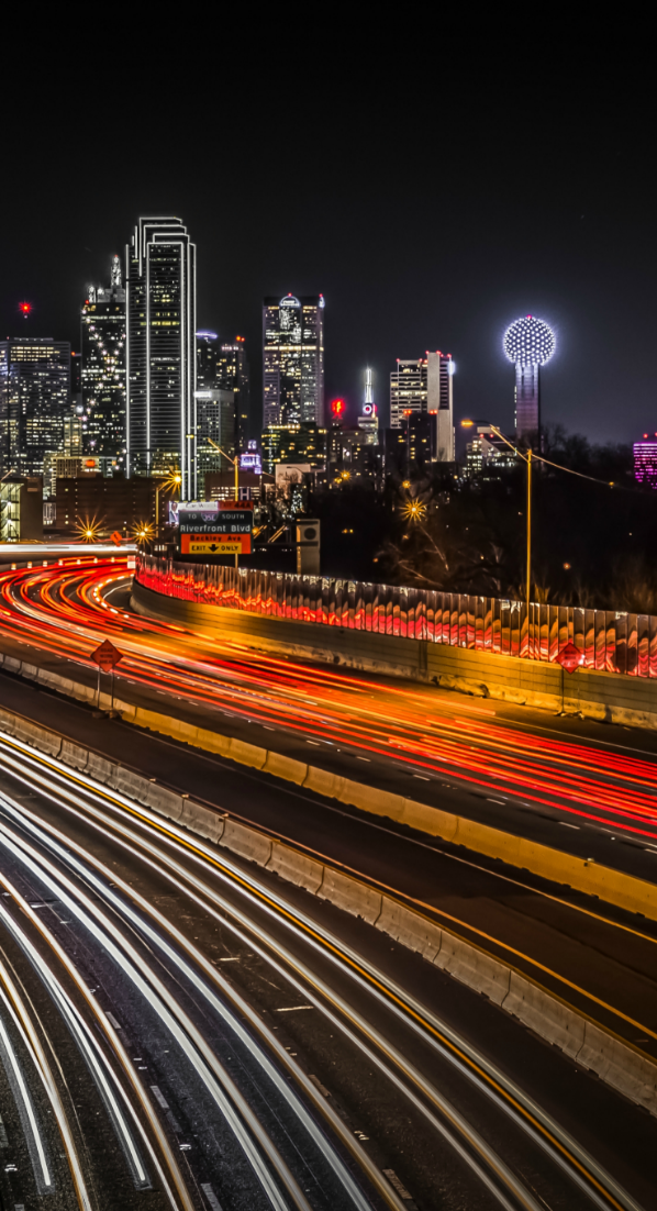 Long-exposure night shot of the Dallas skyline, showing glowing light trails from traffic on a curved highway.