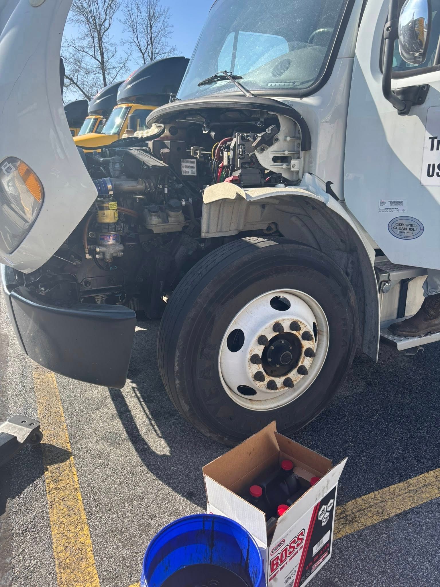 A white commercial truck with its hood open for maintenance, parked on asphalt near a box of oil and a blue bucket.