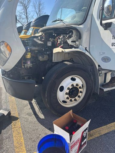 A white commercial truck with its hood open for maintenance, parked on asphalt near a box of oil and a blue bucket.