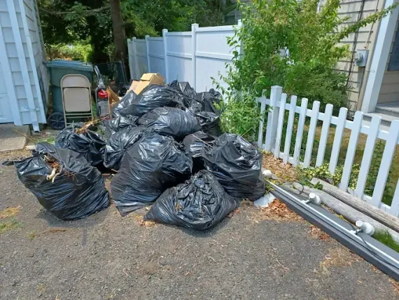Black trash bags piled near a white picket fence, a green trash bin, and a house.