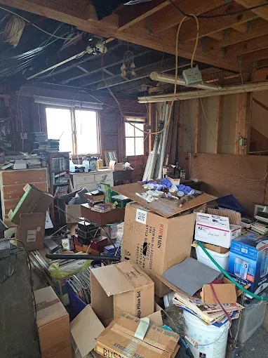 Cluttered garage interior: boxes, debris, and miscellaneous items filling the space. Windows let in natural light.