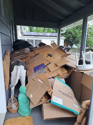 Porch cluttered with cardboard boxes, a trash bag, and decor items, like a statue and a small basket.