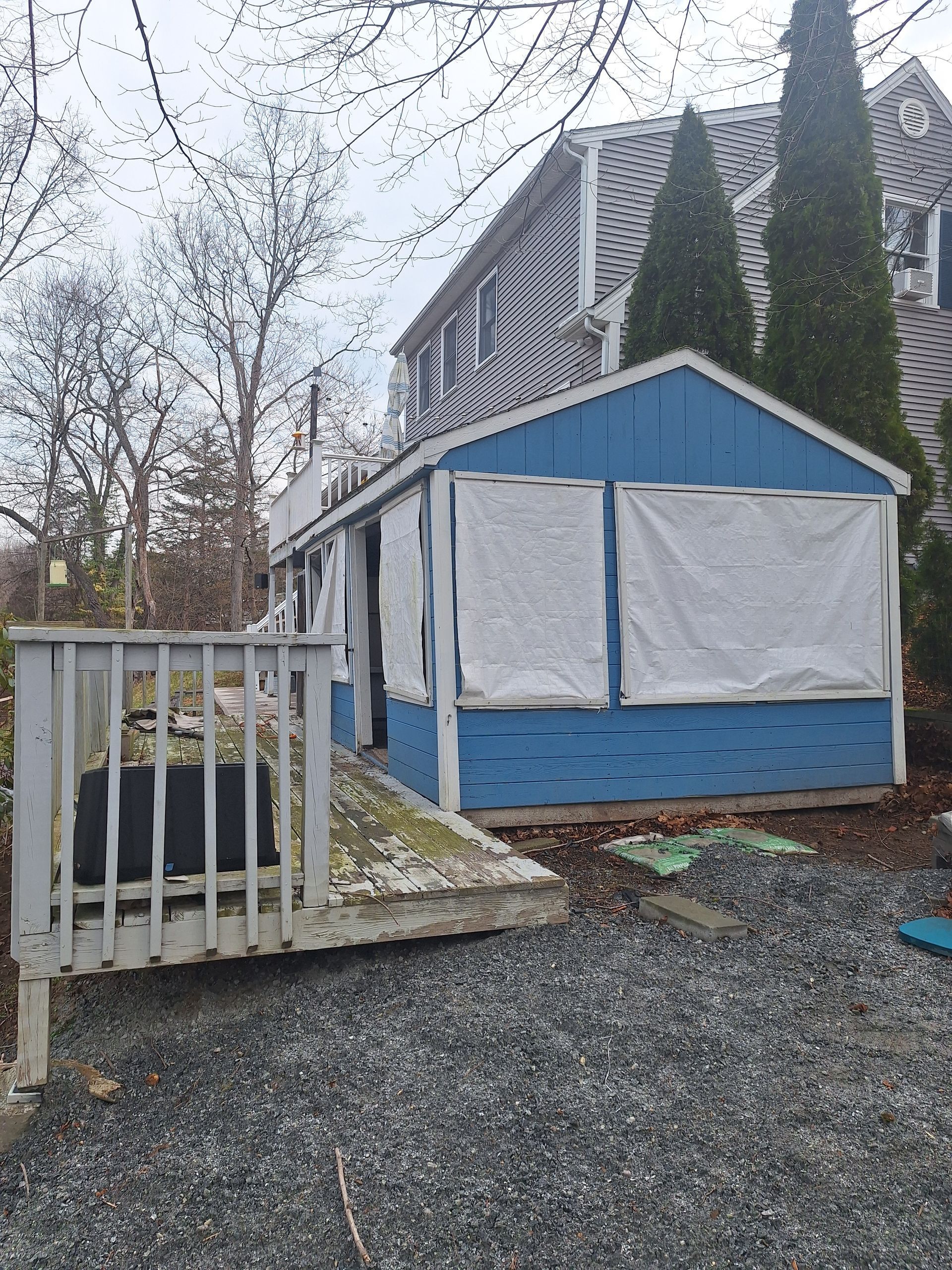 Blue-and-white tarp-covered shed beside a house, with gravel, a fence, and leafless trees