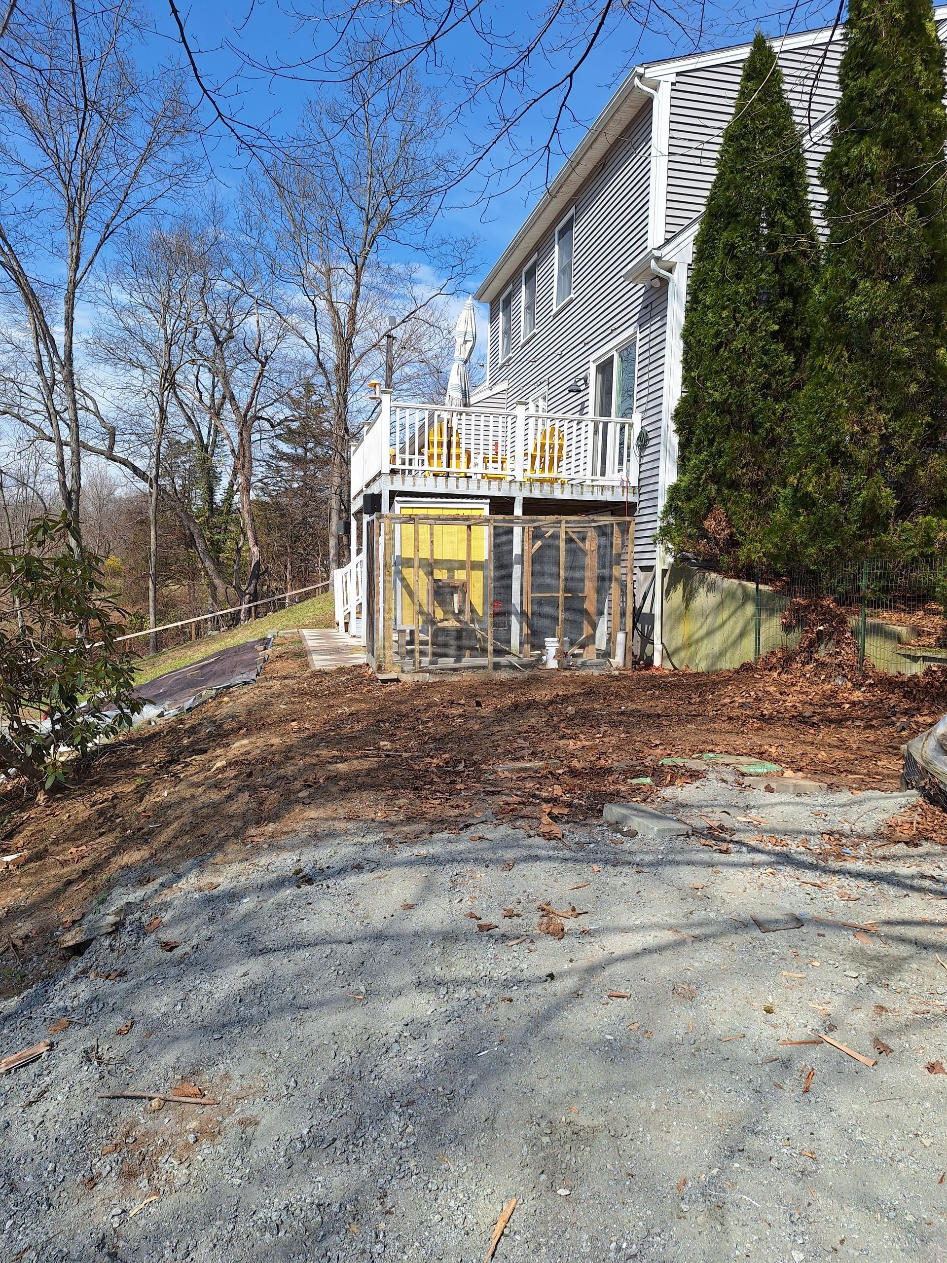 White house with yellow deck beside a gravel driveway, surrounded by trees in late autumn.