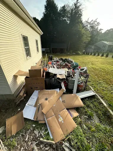 Pile of discarded cardboard boxes and trash next to a beige house, on a grassy lawn with trees in the background.