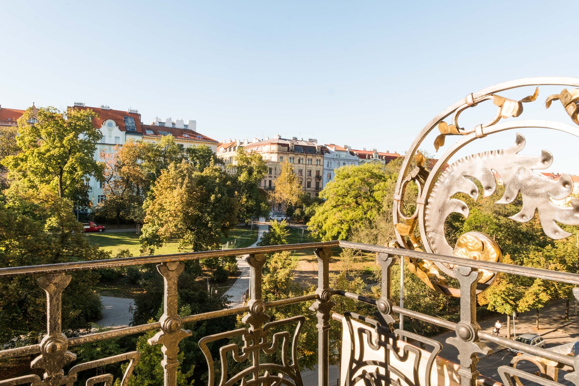 Balkon mit Blick auf einen Park mit Bäumen und Gebäuden; kunstvolles schmiedeeisernes Geländer, sonniger Tag.