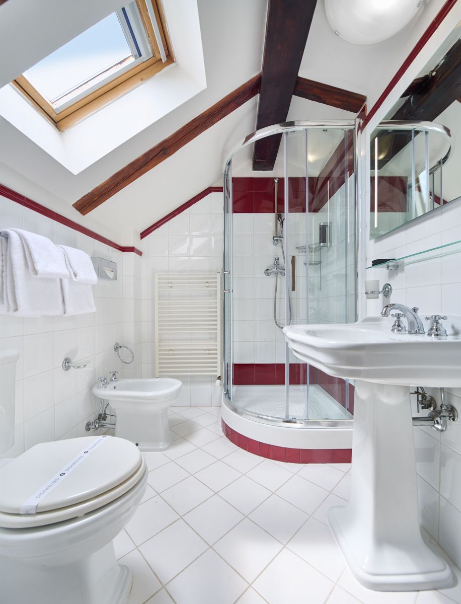 Weißes Badezimmer mit schräger Decke, Oberlicht und roten Akzenten; inklusive Dusche, Toilette, Bidet und Standwaschbecken.