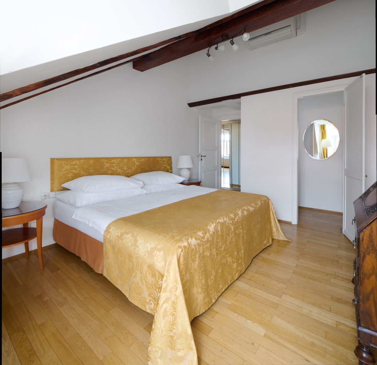 Schlafzimmer mit Bett, Nachttischen und Tür zum Flur, mit goldenen Akzenten. Holzfußboden.