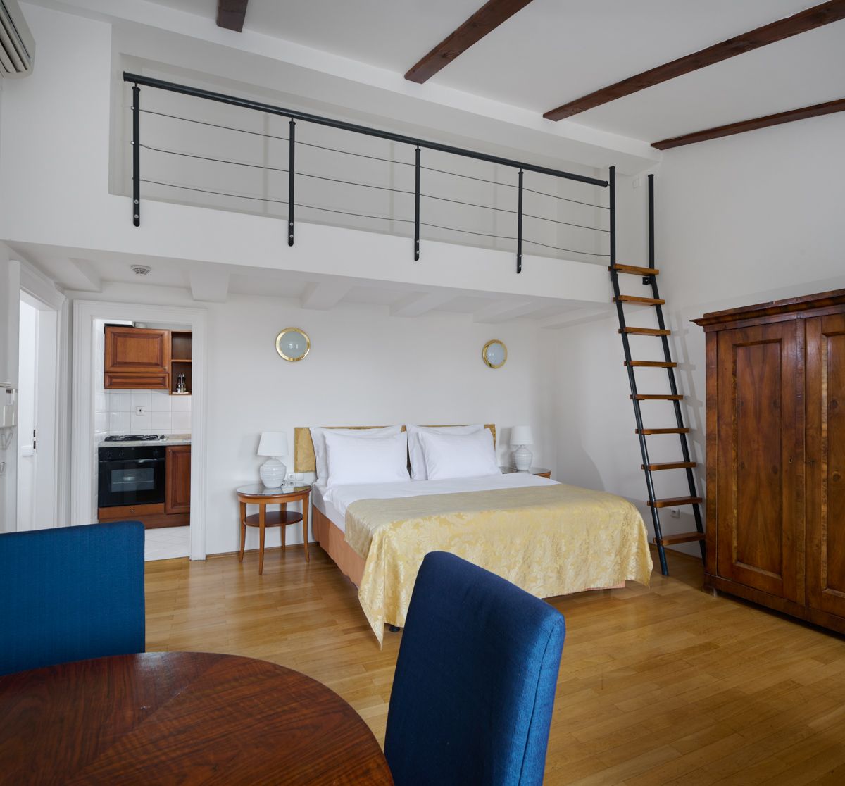 Schlafzimmer mit Bett, kleiner Küche, Galerie und Holzleiter. Das Zimmer hat Holzboden und blaue Stühle.