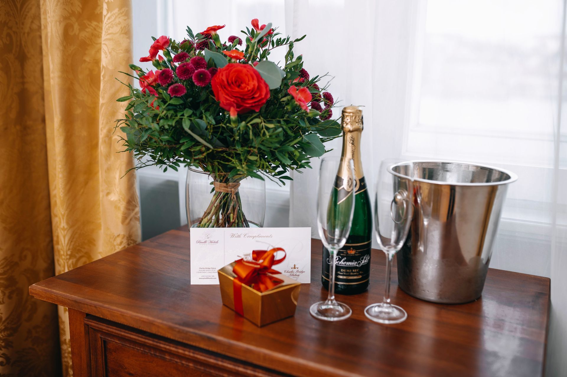 Blumenstrauß, Champagner, Gläser, Geschenk und Eisbehälter auf einem Holztisch in der Nähe eines Fensters.
