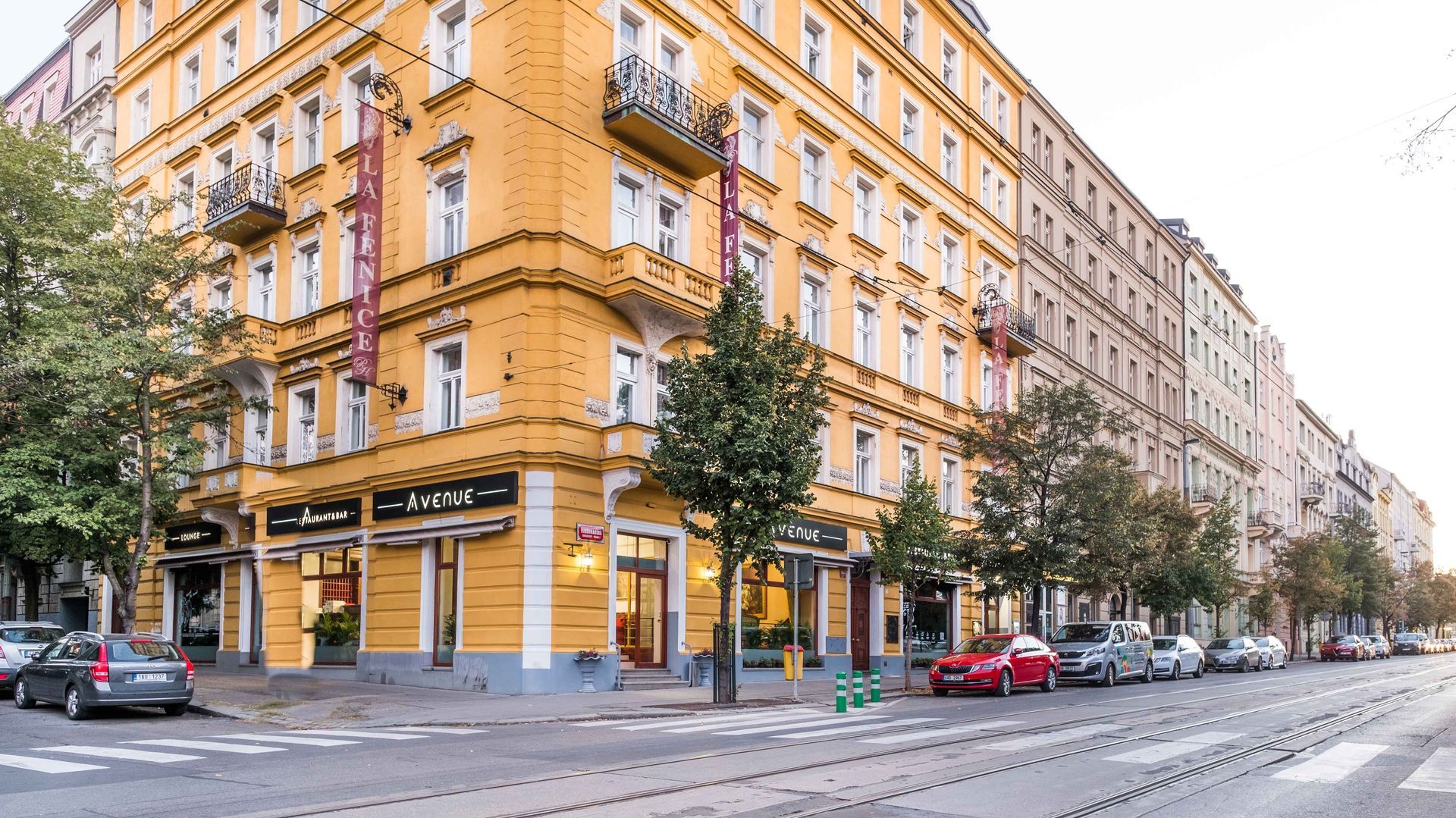 Gelbes Eckgebäude mit Geschäften und Balkonen an einer Stadtstraße; Autos parken am Straßenrand.