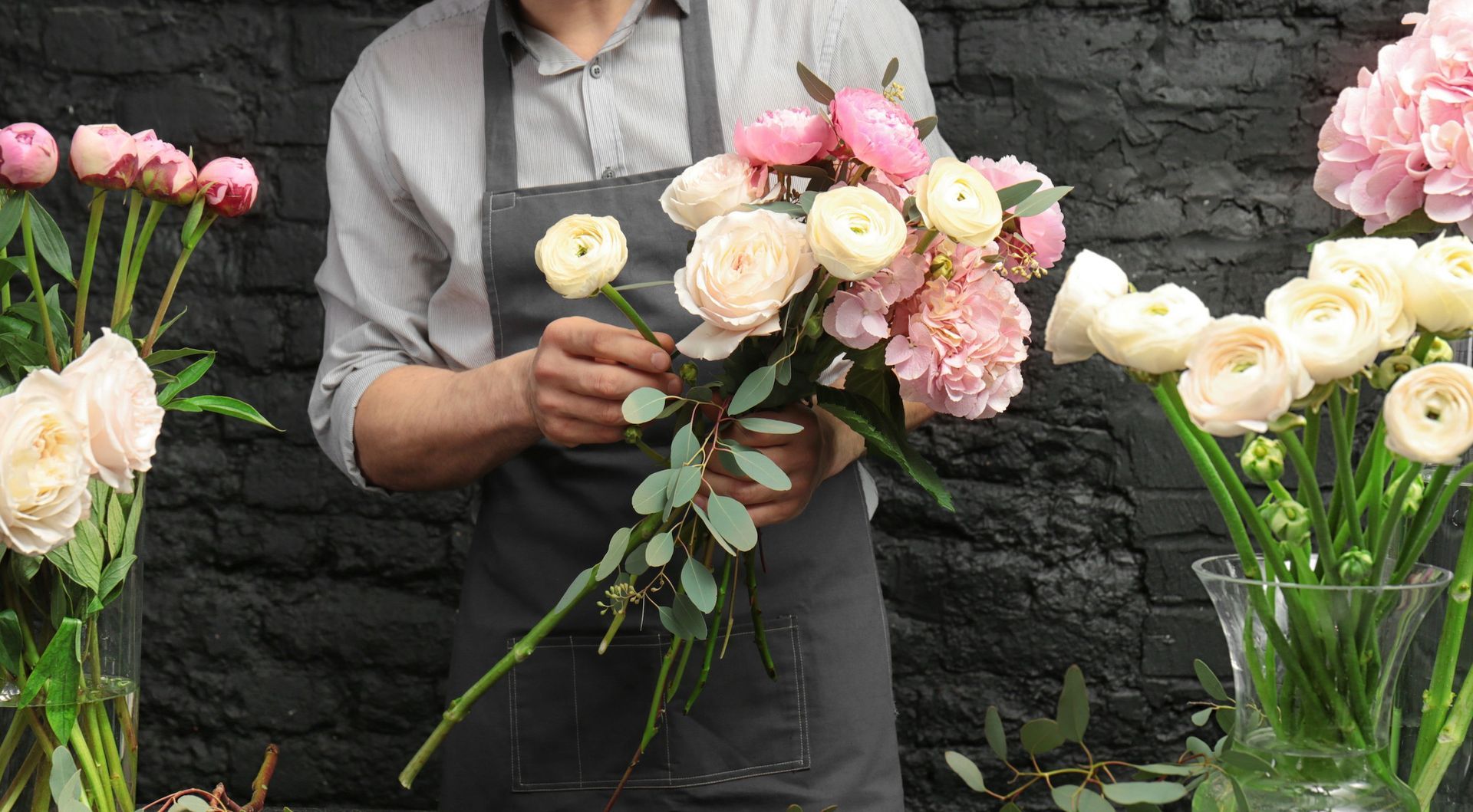 Eine Person, die einen Blumenstrauß aus rosa und cremefarbenen Blumen und Eukalyptus arrangiert und dabei eine Schürze trägt.