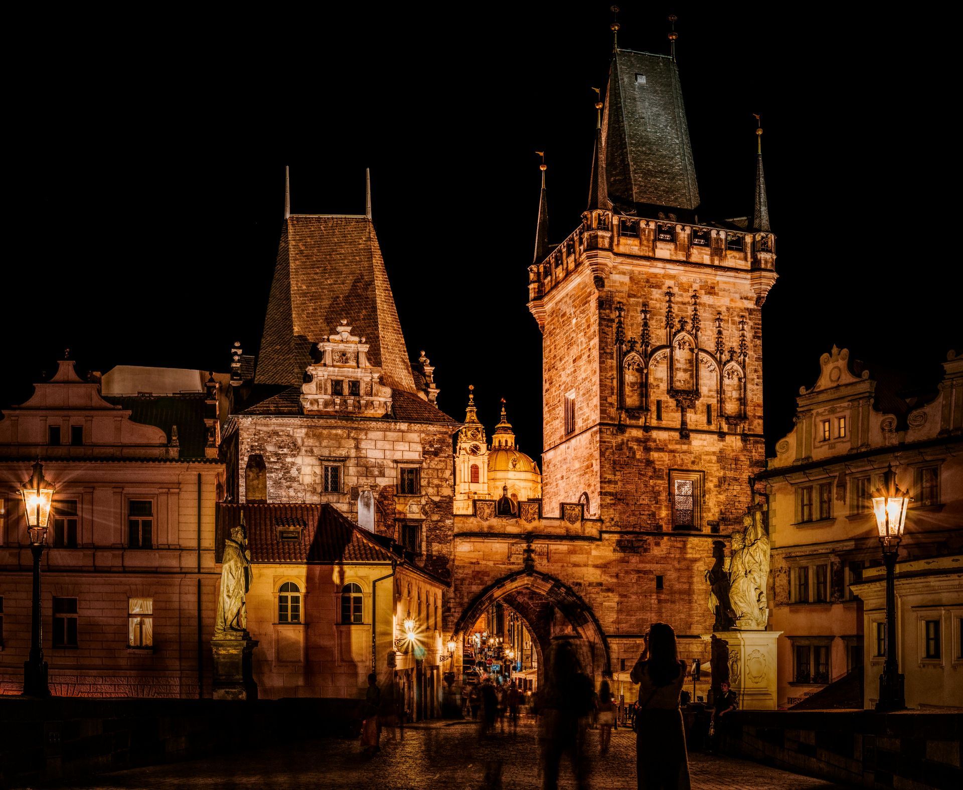 Nachtansicht der Türme der Karlsbrücke in Prag, warm beleuchtet, mit silhouettenhaften Figuren.