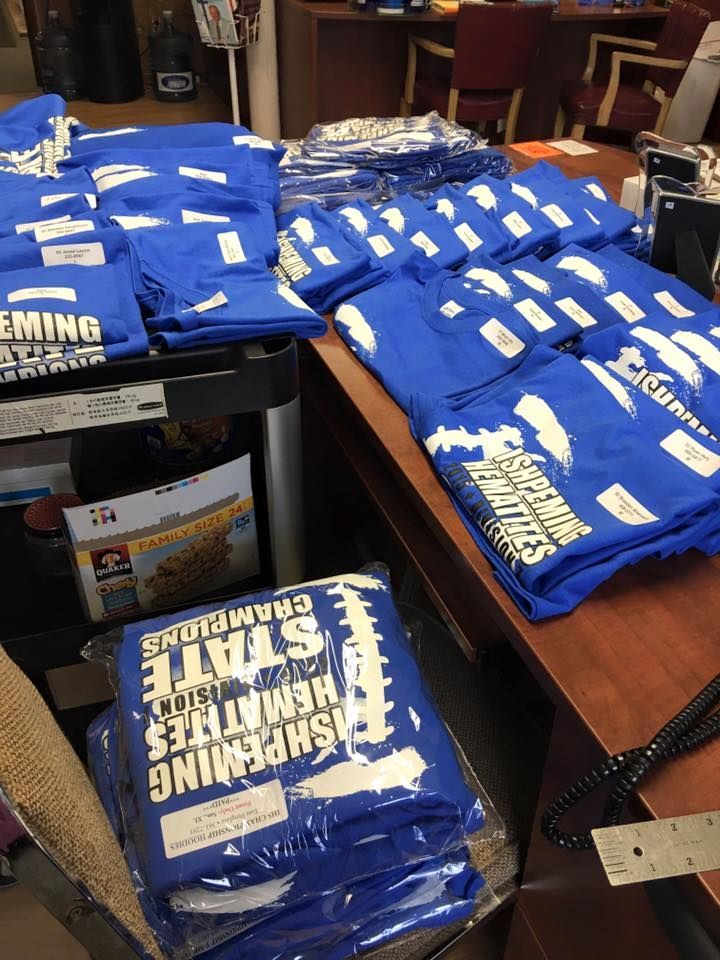 Blue t-shirts stacked on a counter, likely for sale. Shirts have white lettering and a football graphic.