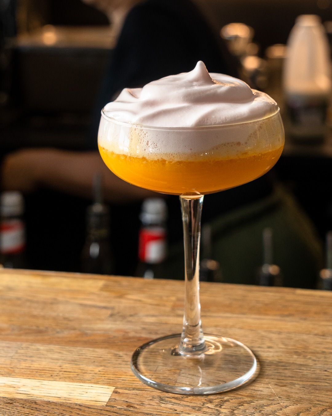 Cocktail in a stemmed glass; orange liquid topped with fluffy white foam, on a wooden bar.