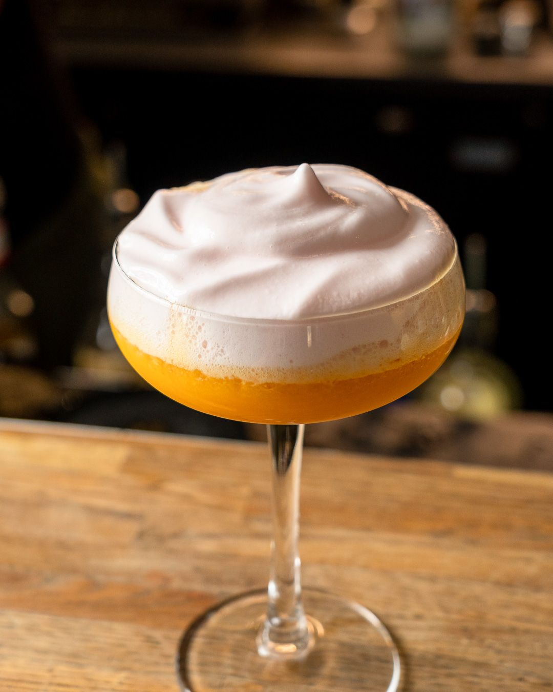 Cocktail in a stemmed glass; orange liquid topped with white foam, set on a wooden bar.