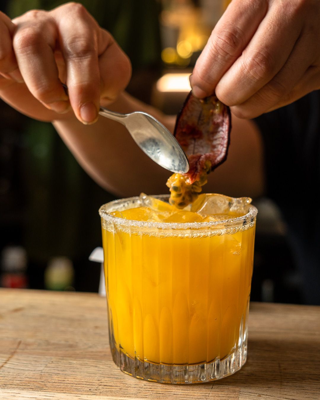 Bartender adding passion fruit pulp to a yellow cocktail in a glass with a sugar-rim, on a wooden surface.