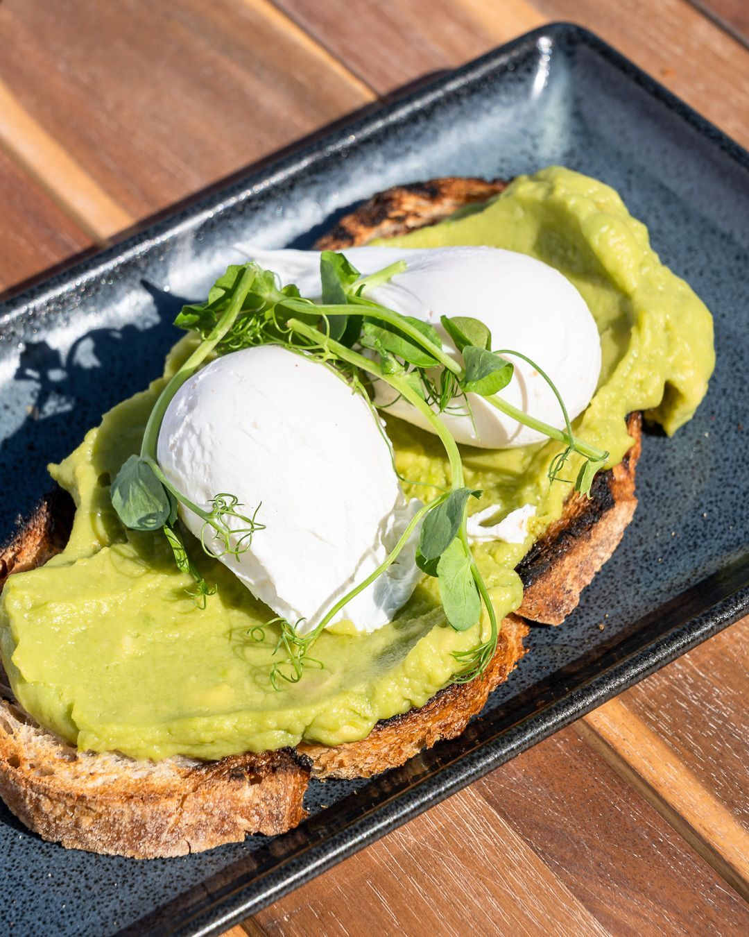 Avocado toast with poached eggs and greens on a dark plate, outdoors.