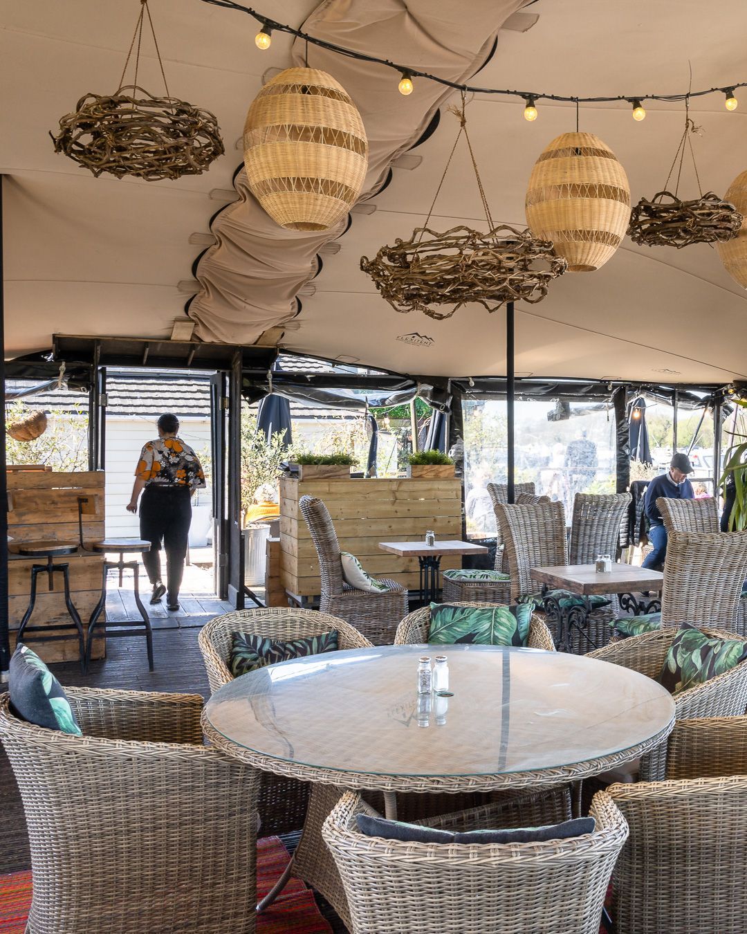 Restaurant patio with wicker furniture, glass-topped tables, woven light fixtures, and a person walking through a doorway.