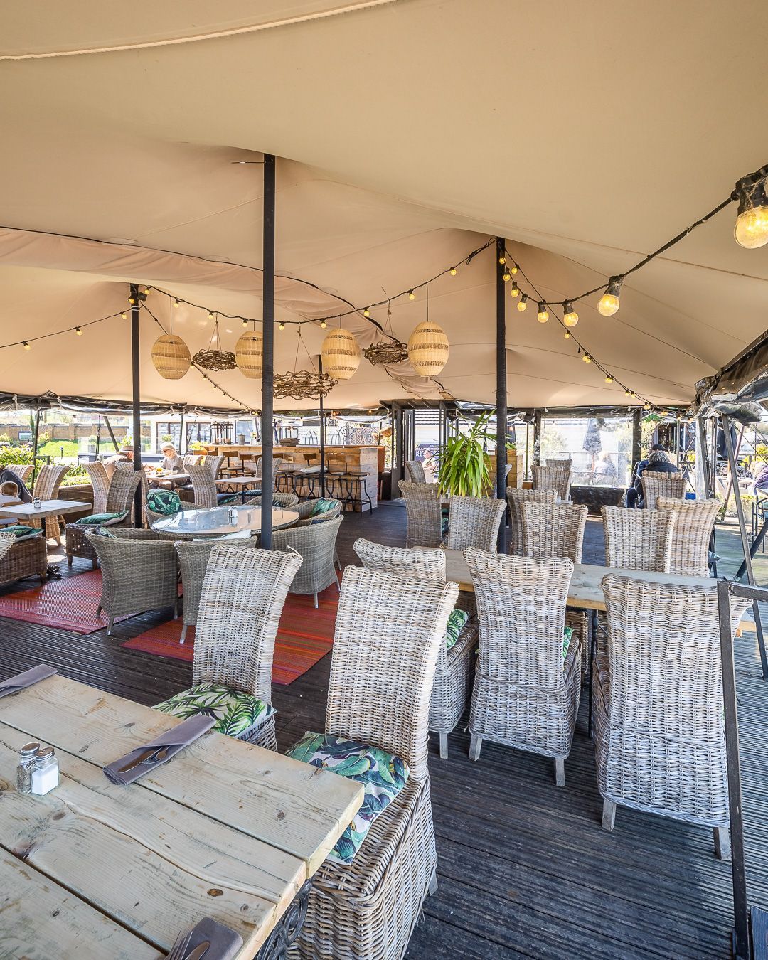 Outdoor restaurant seating area under a cream-colored canopy. Wicker chairs, wooden tables, and string lights visible.