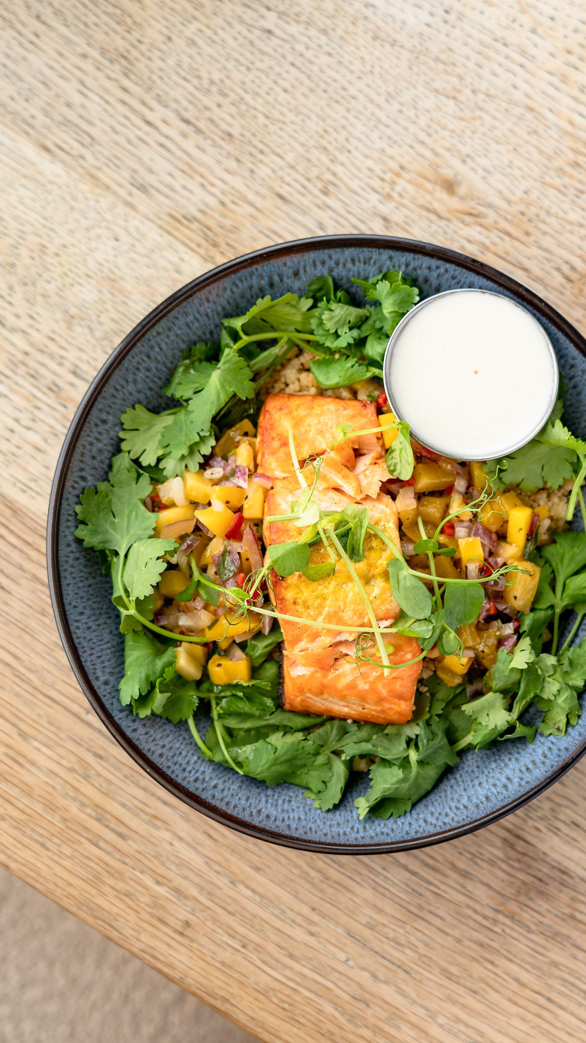 Salmon dish with mango salsa and cilantro in a blue bowl, white sauce on the side.