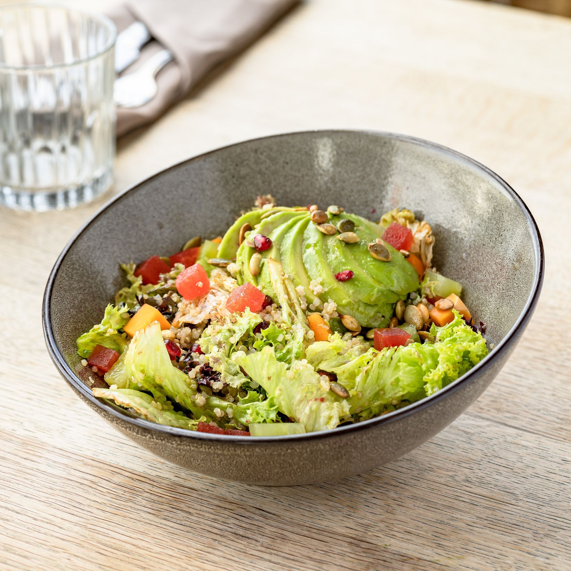 Salad bowl with avocado slices, lettuce, seeds, and chopped vegetables on a wooden table.