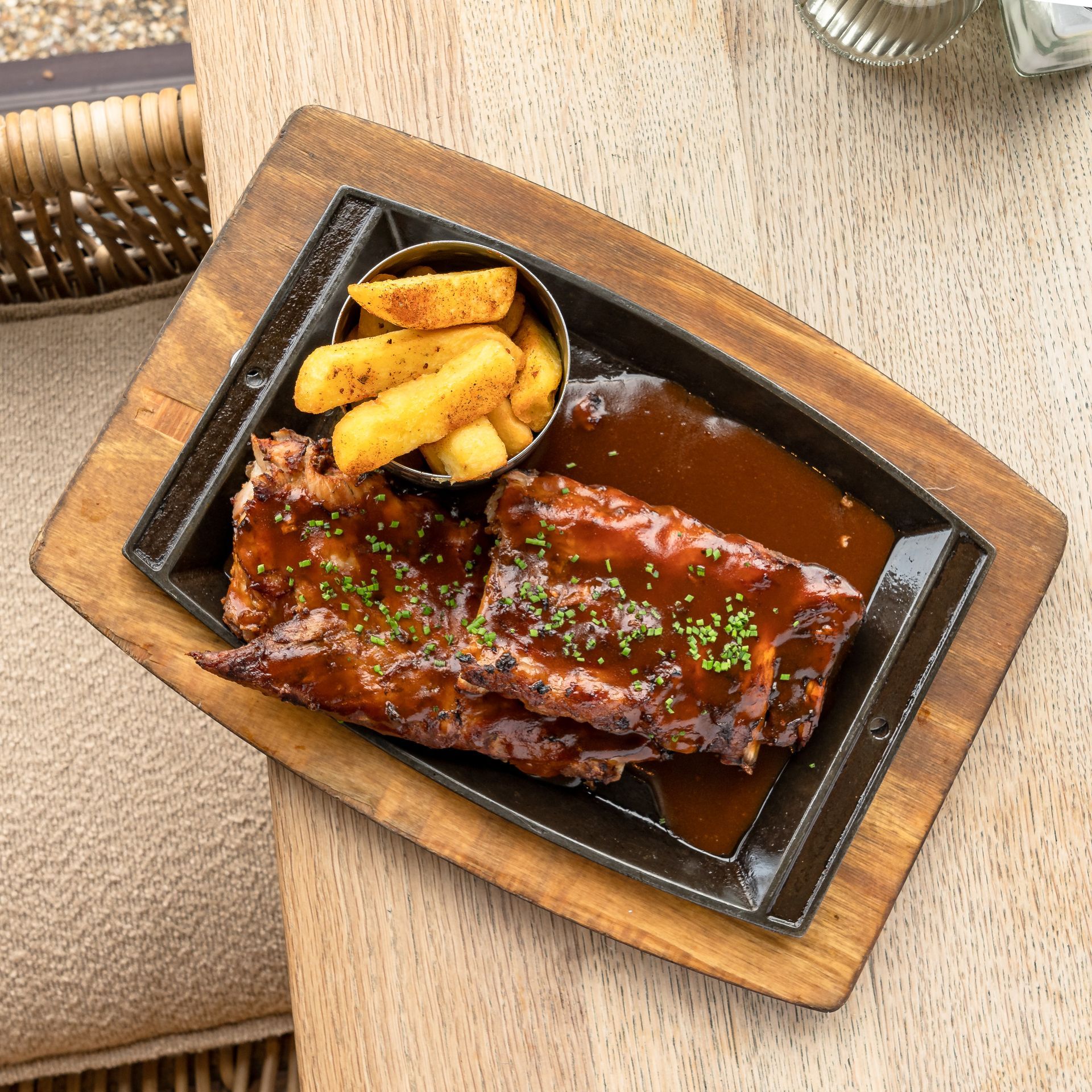Ribs with sauce and fries on a black plate, wooden board, outdoor setting.