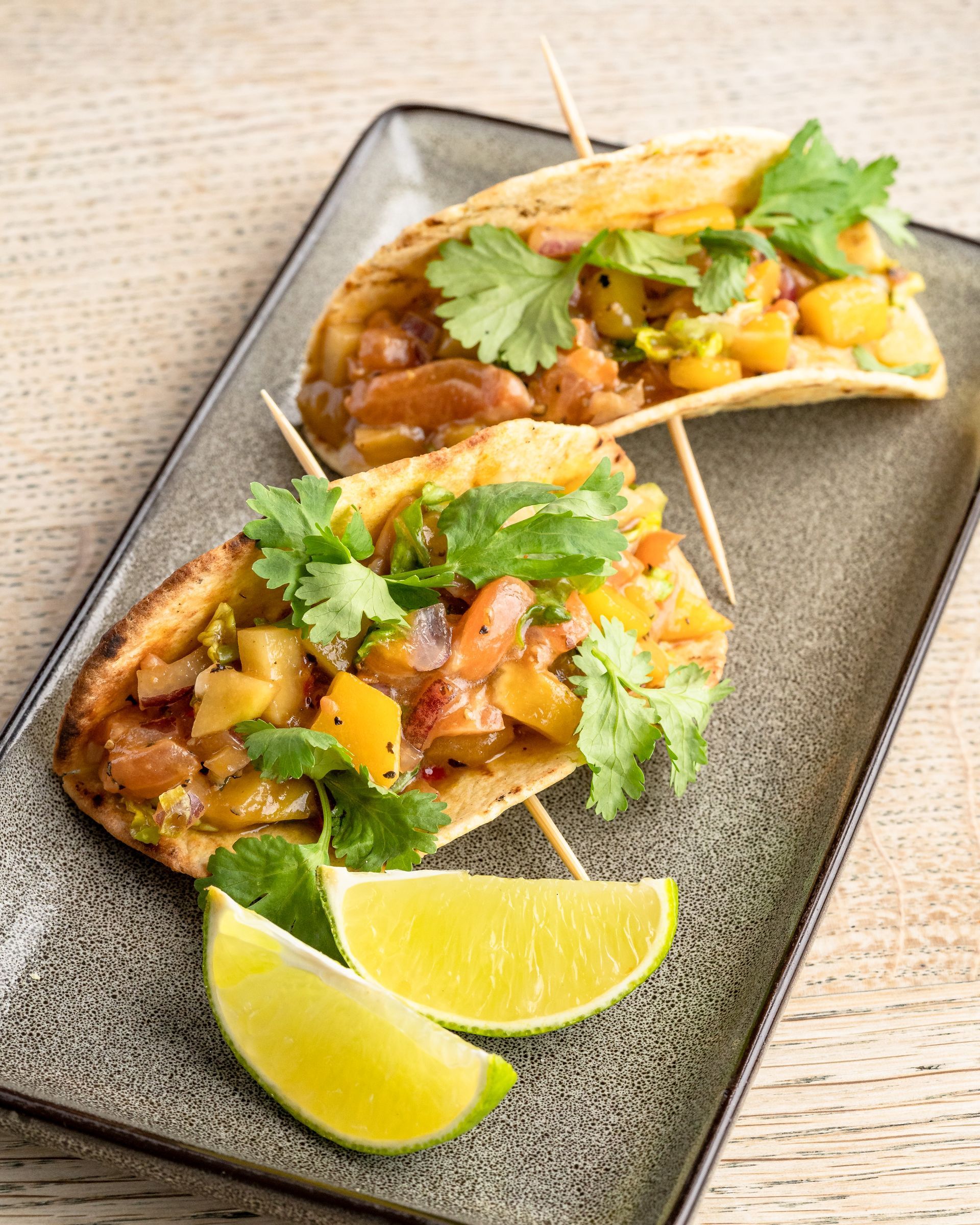 Two open-faced tacos with fruit salsa, cilantro, and lime wedges on a rectangular plate.