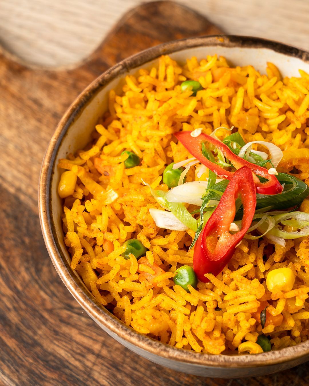 Bowl of yellow rice with vegetables and chili slices on a wooden board.