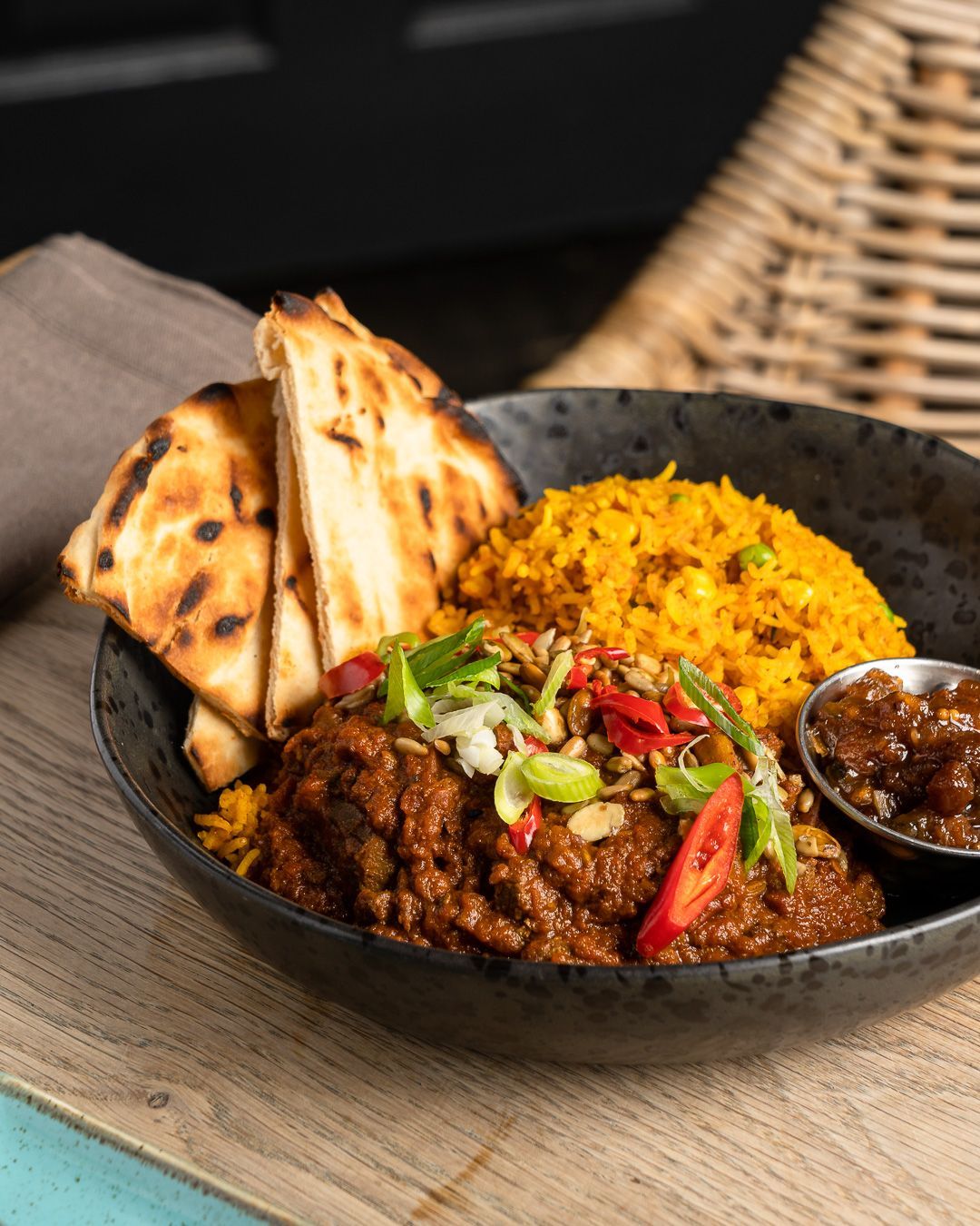 Bowl of curry with rice, naan, and chutney on wooden board.