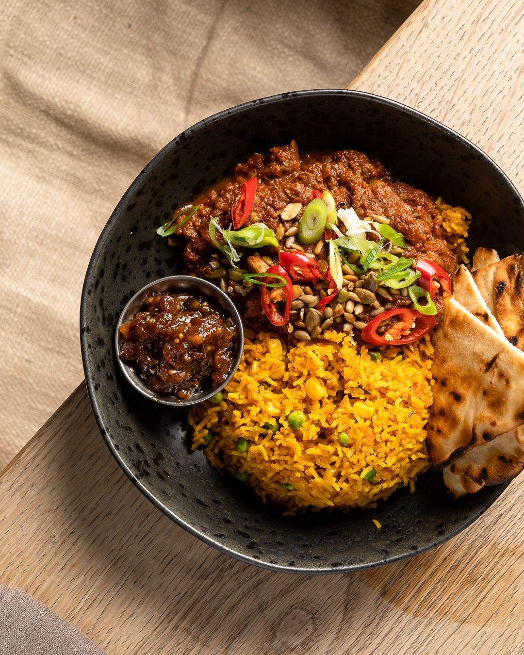 Curried rice, meat, chutney, naan, and toppings in a black bowl on a light-colored surface.