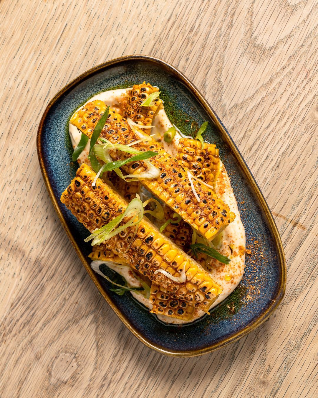 Grilled corn on hummus, garnished with green onions, served on a blue-rimmed plate.