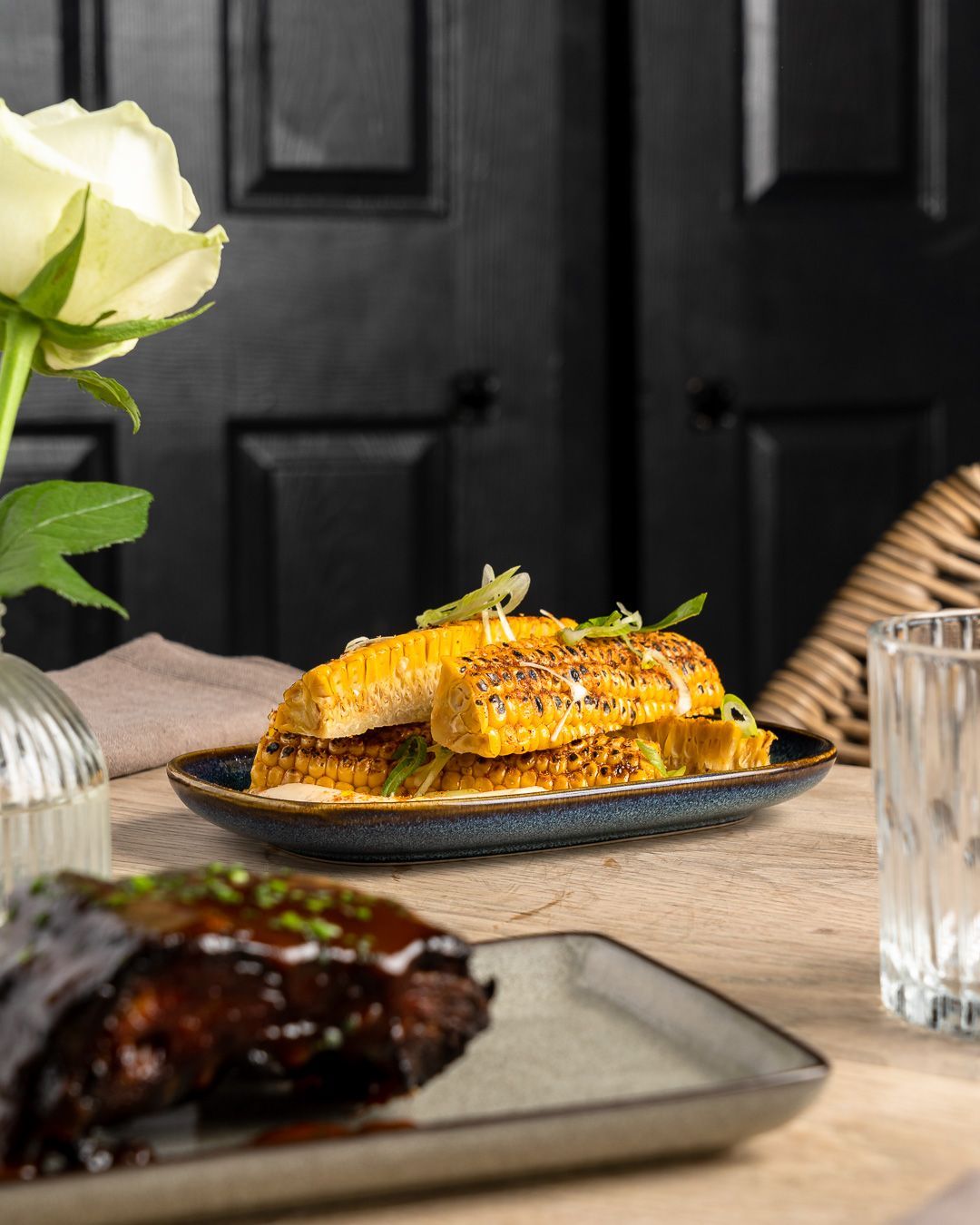 Corn dish on a plate, alongside a plate of meat. Rose in a vase. Wooden table.