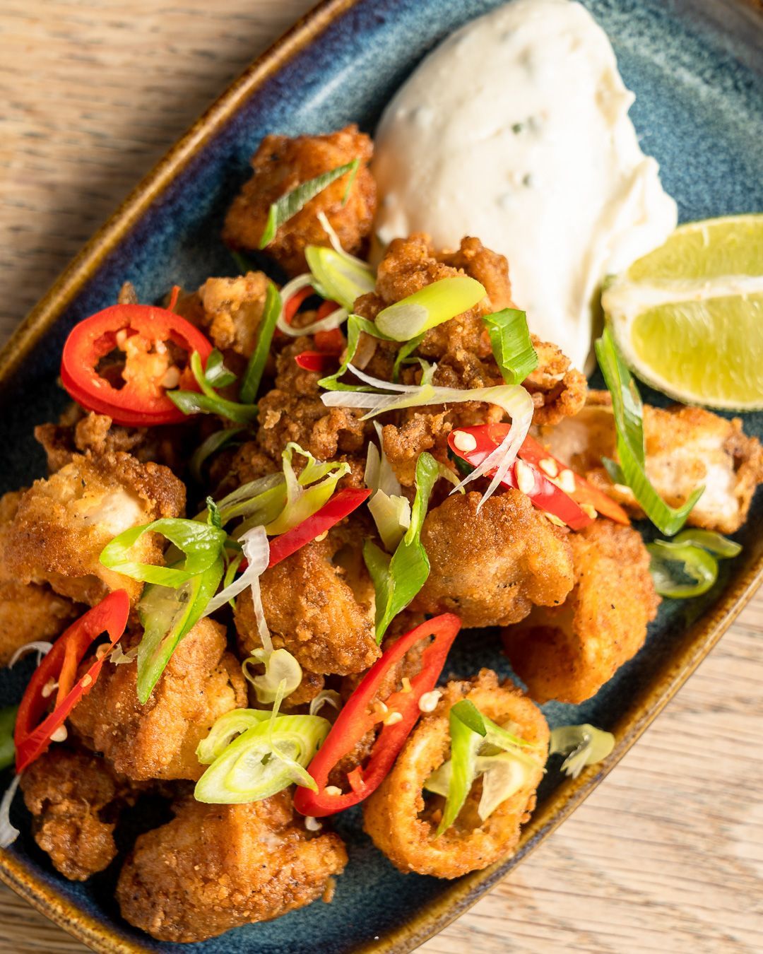 Fried calamari with dipping sauce, red chilies, green onions, and lime wedge on a blue rectangular plate.